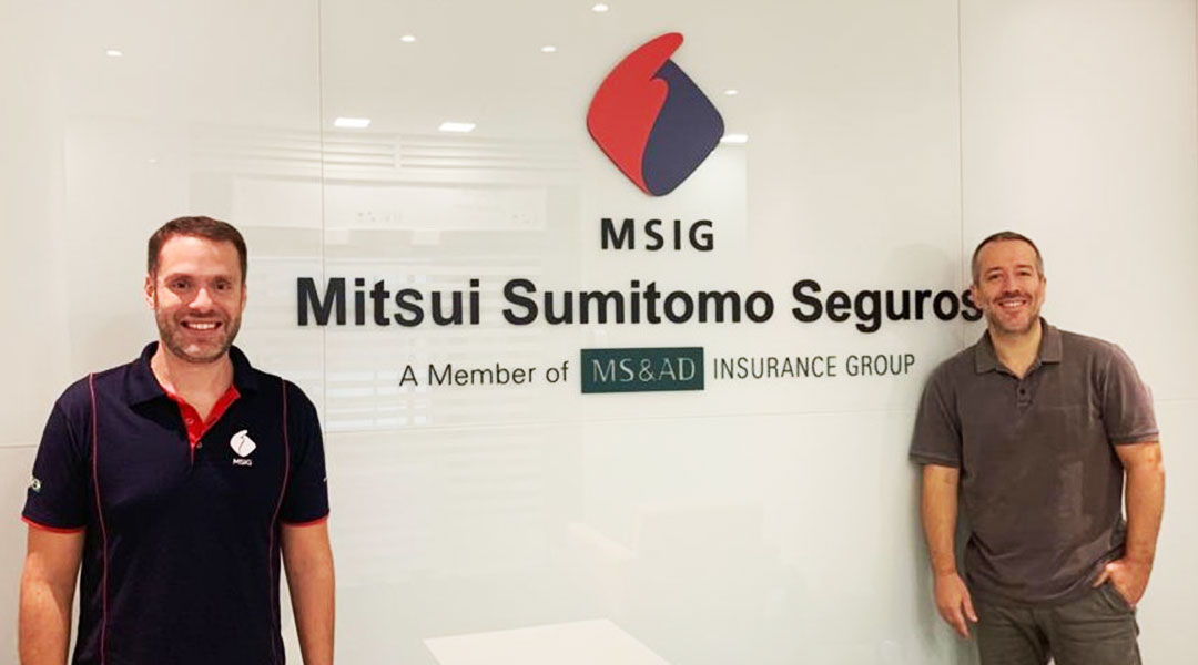 Dois homens sorrindo para a câmera à frente de painel com a marca Mitsui