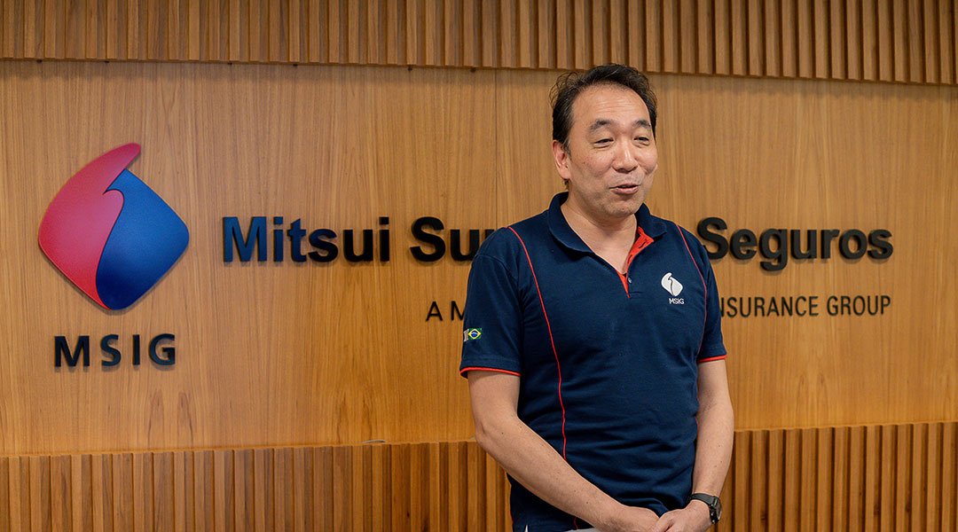 Homem oriental falando à frente de painel de madeira com a marca da Mitsui